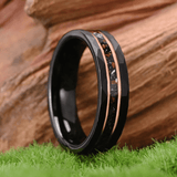 Anneau en tungstène noir 6mm photographié en gros plan, positionné verticalement devant un fond en bois. La surface extérieure est facettée. Une bande centrale texturée avec particules sombres et claires est visible, délimitée par deux lignes métalliques fines de teinte cuivrée.