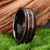Bague circulaire 8 mm en tungstène noir posée verticalement sur une surface verte rappelant de l’herbe. L’anneau présente une surface extérieure facettée. Une bande centrale sombre avec particules visibles traverse la bague, encadrée par deux fines lignes métalliques couleur cuivre. Arrière-plan en bois.