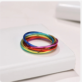 Bague posée sur une surface claire, composée de plusieurs anneaux fins en acier inoxydable entrelacés pour former un cercle unique. Chaque anneau est d’une couleur différente de l’arc-en-ciel, avec une finition lisse et brillante.