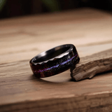 Bague en tungstène noir brillant avec incrustation galaxie irisée bleue et violette, présentée seule sur une surface en bois clair avec un éclairage doux.