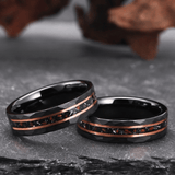 Deux bagues en tungstène noir posées horizontalement sur une surface sombre. Les anneaux présentent une surface extérieure facettée. Une bande centrale texturée avec particules visibles entoure chaque bague, encadrée par deux fines lignes métalliques couleur cuivre. L’intérieur des bagues est lisse.