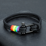 Bracelet composé d’un cordon tressé noir, fermé par une boucle métallique sombre en forme de manille. Près du fermoir, plusieurs ligatures colorées fines disposées à la suite, chacune d’une teinte différente. Les couleurs contrastent légèrement avec le cordon noir, créant un point visuel distinct mais subtil. L’ensemble est présenté dans un coffret noir sur certaines images.