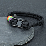 Bracelet composé d’un cordon tressé noir, fermé par une boucle métallique sombre en forme de manille. Près du fermoir, plusieurs ligatures colorées fines disposées à la suite, chacune d’une teinte différente. Les couleurs contrastent légèrement avec le cordon noir, créant un point visuel distinct mais subtil. L’ensemble est présenté dans un coffret noir sur certaines images.