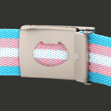 Gros plan sur la boucle métallique rectangulaire d’une ceinture en toile. Une découpe centrale laisse apparaître l 'intérieure de la boucle et la sangle tissée aux couleurs bleu ciel, rose et blanche du drapeau transgenre.