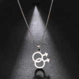 Collier composé d’une fine chaîne métallique portant un pendentif formé de deux cercles entrelacés, chacun prolongé d’une flèche orientée vers l’extérieur. Le pendentif représente le symbole masculin doublé. L’ensemble existe en finition dorée ou argentée. Le pendentif forme une silhouette nette, géométrique, avec des lignes lisses et une surface uniforme. La lumière glisse sur le métal et crée une ombre ovale sous le pendentif, soulignant la forme des deux cercles reliés.