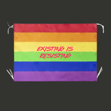 Drapeau rectangulaire avec bandes horizontales rouge, orange, jaune, vert, bleu et violet formant le drapeau arc-en-ciel Pride. Au centre apparaît le texte rose vif "Existing Is Resisting" écrit dans une typographie manuscrite dynamique. Des bandes de fixation blanches sont visibles aux coins.