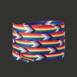 Paire de lacets plats enroulés. Les lacets présentent des bandes multicolores horizontales correspondant aux couleurs du drapeau Pride. Ce modèle montre un motif en chevrons avec des bandes supplémentaires. Les couleurs sont vives et contrastées. Les extrémités sont rigides. Présentation sur fond gris sans texte intégré