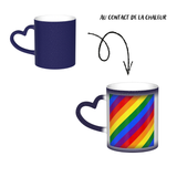 Mug en céramique avec poignée en forme de cœur. À froid, l’extérieur est uniformément coloré (noir, bleu ou rouge) avec de petits points blancs. Au contact de la chaleur, le centre devient blanc et révèle un grand rectangle composé de bandes diagonales arc-en-ciel : rouge, orange, jaune, vert, bleu et violet, orientées du coin supérieur gauche vers le coin inférieur droit. Le contraste reste visible entre la zone éclaircie et la couleur d’origine du mug.