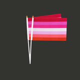 Deux petits drapeaux lesbiens montés sur tiges blanches avec embouts dorés, présentés légèrement inclinés sur fond gris. 