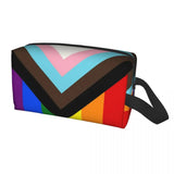 Trousse rectangulaire à fermeture éclair, motif “Progress Pride”. Sur le corps de la trousse figurent des bandes verticales aux couleurs de l’arc-en-ciel (rouge, orange, jaune, vert, bleu, violet), ainsi qu’un chevron sur le dessus, formé de bandes noires, marron, bleu clair, rose et blanc. L’ensemble rappelle le drapeau de la fierté LGBTQIA+, symbolisant la diversité et l’inclusion. La trousse est de forme allongée, avec une petite sangle noire sur le côté.