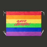 Drapeau rectangulaire aux bandes horizontales arc-en-ciel rouge, orange, jaune, vert, bleu et violet. Au centre apparaît le texte rose vif "Queer Liberation" écrit dans une typographie manuscrite dynamique. Des attaches blanches sont visibles aux coins pour suspendre le drapeau.