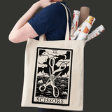 Sac tote bag en tissu écru porté à l’épaule. Sur la face avant, une illustration noire encadrée représente une paire de ciseaux stylisés au-dessus de vagues et de nuages, avec le mot “SCISSORS.” en bas. Le visuel fait référence à un symbole de la culture lesbienne WLW.