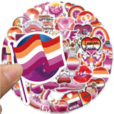 Photo d’un sticker représentant un drapeau lesbien stylisé, tenu entre deux doigts, avec un fond flou composé d’autres stickers assortis.