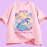  T-shirt unisexe décliné en plusieurs couleurs. Sur le devant, une grande illustration centrale : un dauphin bleu-violet sautant hors de l’eau. Derrière lui, un arc de couleurs et un ciel jaune et violet. Autour, fleurs roses et petits cœurs colorés. Au-dessus : le mot “RAGE” en lettres majuscules rose foncé. En dessous : “CONSUMES ME” dans la même typographie. Le visuel occupe une zone rectangulaire centrée sur le torse. 