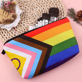 Trousse rectangulaire de petite taille, imprimée du motif “Pride Progress”. Le motif combine des bandes horizontales rouge, orange, jaune, vert, bleu, violet, et à gauche une chevron verticale avec rose, bleu clair, blanc, marron, noir. La trousse est ouverte — la doublure intérieure est d’un tissu sombre uni. Une fermeture éclair traverse la largeur supérieure. La trousse apparaît vide ou très légèrement ouverte, montrant l’intérieur sombre, sans objets à l’intérieur visibles.