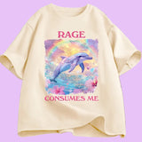T-shirt unisexe décliné en plusieurs couleurs. Sur le devant, une grande illustration centrale : un dauphin bleu-violet sautant hors de l’eau. Derrière lui, un arc de couleurs et un ciel jaune et violet. Autour, fleurs roses et petits cœurs colorés. Au-dessus : le mot “RAGE” en lettres majuscules rose foncé. En dessous : “CONSUMES ME” dans la même typographie. Le visuel occupe une zone rectangulaire centrée sur le torse.