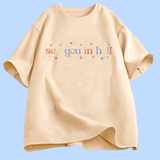 T-shirt à manches courtes présenté à plat. Au centre, le texte « see you in hell » écrit en lettres minuscules, avec un dégradé de couleurs. Autour du texte, de petites étoiles décoratives multicolores. Le motif est centré sur la poitrine. Le t-shirt existe en plusieurs couleurs unies. Aucun autre texte ou image n’est présent.