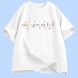 T-shirt à manches courtes présenté à plat. Au centre, le texte « see you in hell » écrit en lettres minuscules, avec un dégradé de couleurs. Autour du texte, de petites étoiles décoratives multicolores. Le motif est centré sur la poitrine. Le t-shirt existe en plusieurs couleurs unies. Aucun autre texte ou image n’est présent.