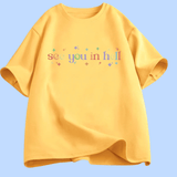 T-shirt à manches courtes présenté à plat. Au centre, le texte « see you in hell » écrit en lettres minuscules, avec un dégradé de couleurs. Autour du texte, de petites étoiles décoratives multicolores. Le motif est centré sur la poitrine. Le t-shirt existe en plusieurs couleurs unies. Aucun autre texte ou image n’est présent.