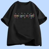 T-shirt à manches courtes présenté à plat. Au centre, le texte « see you in hell » écrit en lettres minuscules, avec un dégradé de couleurs. Autour du texte, de petites étoiles décoratives multicolores. Le motif est centré sur la poitrine. Le t-shirt existe en plusieurs couleurs unies. Aucun autre texte ou image n’est présent.