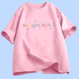 T-shirt à manches courtes présenté à plat. Au centre, le texte « see you in hell » écrit en lettres minuscules, avec un dégradé de couleurs. Autour du texte, de petites étoiles décoratives multicolores. Le motif est centré sur la poitrine. Le t-shirt existe en plusieurs couleurs unies. Aucun autre texte ou image n’est présent.
